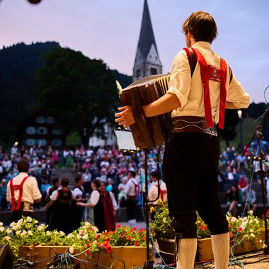 A musician plays an accordion in front of a large crowd. The atmosphere is festive, surrounded by a beautiful natural landscape. | © Kleinwalsertal Tourismus | Oliver Farys