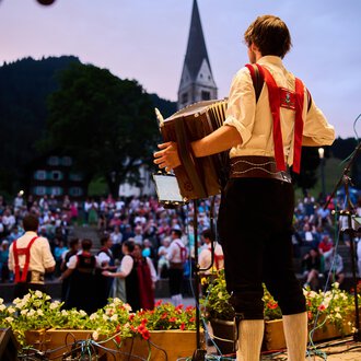A musician plays an accordion in front of a large crowd. The atmosphere is festive, surrounded by a beautiful natural landscape. | © Kleinwalsertal Tourismus | Oliver Farys