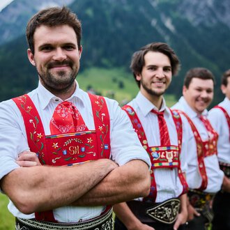 Four men in traditional attire stand in front of a picturesque mountain landscape. They smile friendly at the camera. | © Kleinwalsertal Tourismus | Oliver Farys
