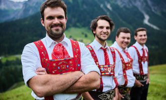 Four men in traditional attire stand in front of a picturesque mountain landscape. They smile friendly at the camera. | © Kleinwalsertal Tourismus | Oliver Farys