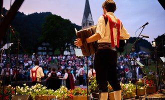 Ein Musiker spielt auf einem Akkordeon vor einer großen Menschenmenge. Im Hintergrund ist eine Kirche und eine malerische Landschaft zu sehen. | © Kleinwalsertal Tourismus | Oliver Farys