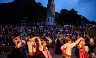 Eine Menschenmenge versammelt sich vor einer Kirche, während Tänzer in traditionellen Kostümen eine Aufführung zeigen. Die Stimmung ist festlich und lebhaft bei Abenddämmerung. | © Kleinwalsertal Tourismus | Oliver Farys