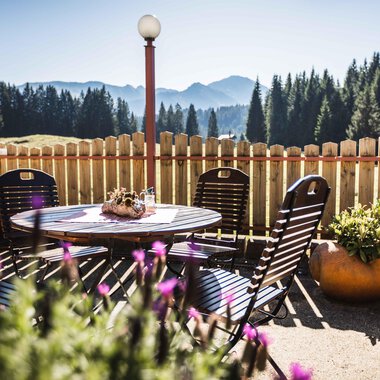 A cozy terrace with a round table and chairs. In the background, mountains and coniferous trees are visible, and the sun is shining. | © Alpenhotel DAS KÜREN