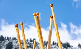 A group of people is playing alphorns on a terrace. In the background, sunny mountains and a clear blue sky can be seen. | © Kleinwalsertal Tourismus | Frank Drechsel
