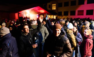 Eine lebhafte Menschenmenge feiert nachts unter einem zeltartigen Pavillon. Viele Personen tragen warme Kleidung und scheinen Spaß zu haben.