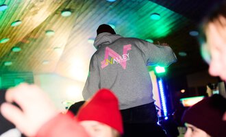 Eine Person steht auf einer Plattform und trägt einen grauen Kapuzenpullover mit dem Schriftzug "Valley". Im Hintergrund sind bunte Lichter und der fröhliche Trubel einer Feier zu sehen.