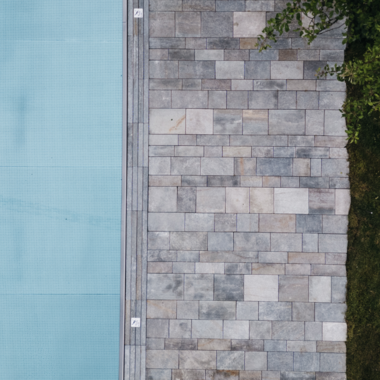 Ein modern gestalteter Außenbereich mit einem blauen Pool und einem Steinweg. Rechts daneben liegt ein grüner Rasen und etwas Laub. | © Naturhotel Chesa Valisa