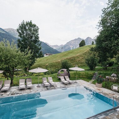 Ein schöner Pool mit Liegen und Sonnenschirmen in einer malerischen Umgebung. Im Hintergrund sind grüne Hügel und Berge zu sehen.