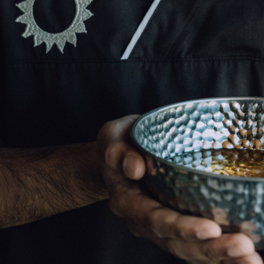 A person is holding a decorative, forged bowl with a golden liquid. The background is dark and focuses on the bowl. | © Naturhotel Chesa Valisa