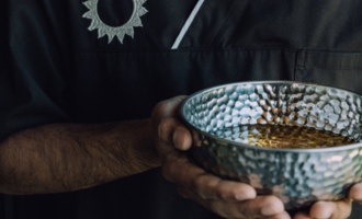 A person is holding a decorative, forged bowl with a golden liquid. The background is dark and focuses on the bowl. | © Naturhotel Chesa Valisa