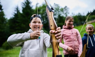 Two people in the woods shooting arrows. The woman is wearing a colorful sweater and aims with a bow, while the man stands behind her in a green T-shirt. | © Kleinwalsertal Tourismus eGen - Fotograf: Oliver Farys