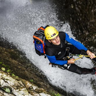 A man is climbing a steep rock wall in a waterfall. He is wearing a helmet and climbing gear. | © Bergschule Kleinwalsertal | Dominik Berchtold