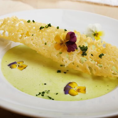 An artistically arranged plate with a crispy waffle structure and a creamy, green soup. Colorful edible flowers add a special touch to the dish.