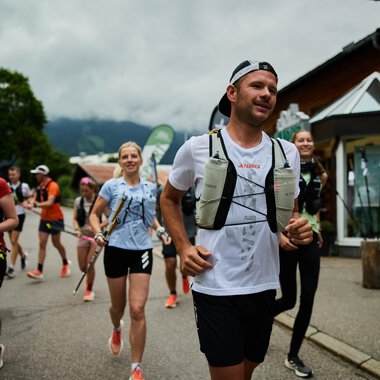 A group of runners is jogging on a road in nature. In the background, trees and a building are visible, and the sky is cloudy. | © Kleinwalsertal Tourismus | Oliver Farys