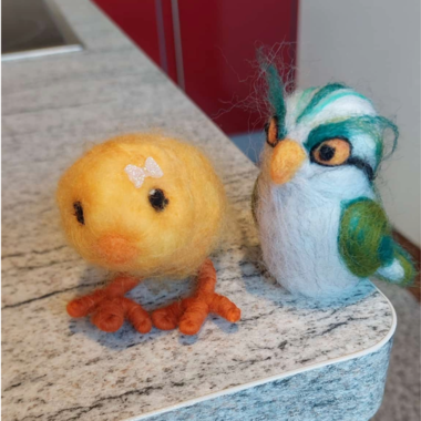 Ein gelbes, buntes Küken und ein gefilzter Vogel stehen nebeneinander auf einer grauen Oberfläche. Beide Figuren sind liebevoll gestaltet und zeigen lebendige Farben. | © Maria Möckel, Der Kleinwalsertaler Rosenhof