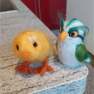 Ein gelbes, buntes Küken und ein gefilzter Vogel stehen nebeneinander auf einer grauen Oberfläche. Beide Figuren sind liebevoll gestaltet und zeigen lebendige Farben. | © Maria Möckel, Der Kleinwalsertaler Rosenhof