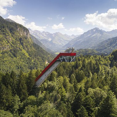 Eine beeindruckende Aussichtsplattform, die über die Bäume ragt, umgeben von majestätischen Bergen und dichten Wäldern. Der Himmel ist klar und zeigt einige Wolken.