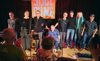 Eine Band steht auf der Bühne und lächelt in die Kamera. Im Hintergrund ist ein leuchtendes Banner mit der Aufschrift "SOUL" zu sehen. | © Soulmine