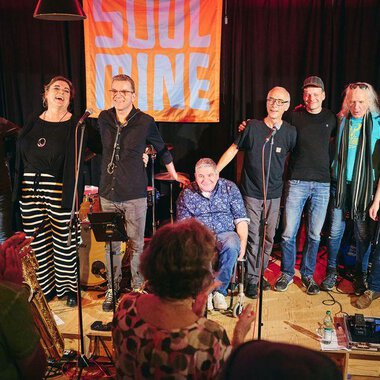 Eine Band steht auf der Bühne und lächelt in die Kamera. Im Hintergrund ist ein leuchtendes Banner mit der Aufschrift "SOUL" zu sehen. | © Soulmine