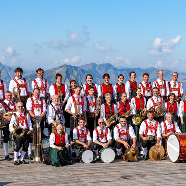 A large brass band in traditional attire stands in front of an impressive mountain backdrop. In the background, the mountains can be seen under a blue sky. | © Trachtenkapelle Riezlern