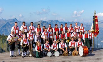A large brass band in traditional attire stands in front of an impressive mountain backdrop. In the background, the mountains can be seen under a blue sky. | © Trachtenkapelle Riezlern