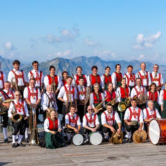 Eine große Brassband mit traditioneller Tracht steht vor einer beeindruckenden Bergkulisse. Im Hintergrund sind die Berge unter einem blauen Himmel zu sehen. | © Trachtenkapelle Riezlern