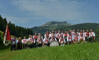 Eine Gruppe von Musikern steht in einem grünen Feld mit Bergen im Hintergrund. Sie tragen traditionelle Kleidung und halten Instrumente in den Händen. | © Musikverein d´Hirschegger