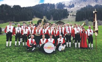 Eine Blaskapelle in traditionellen Trachten steht auf einer grünen Wiese. Im Hintergrund sind Berge und ein klarer Himmel zu sehen. | © Trachtenkapelle Harmonie Mittelberg