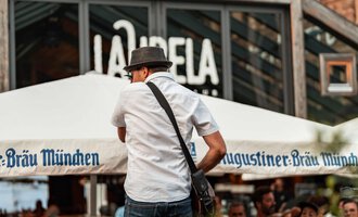 A lively street scene with a crowd watching a performer. In the background, the restaurant "La Capela" can be seen. | © Auenhütte | Basti Heckl
