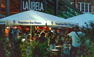 A lively atmosphere under a tent with the inscription "Augustiner-Bräu München". People enjoy music and community in a bustling outdoor area. | © Auenhütte | Basti Heckl