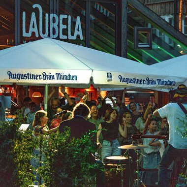 A lively atmosphere under a tent with the inscription "Augustiner-Bräu München". People enjoy music and community in a bustling outdoor area. | © Auenhütte | Basti Heckl