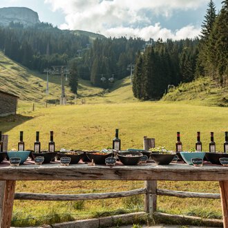 A beautiful outdoor table set with wine bottles and various dishes. In the background, gentle hills and trees are visible. | © Auenhütte | Basti Heckl
