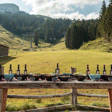 A beautiful outdoor table set with wine bottles and various dishes. In the background, gentle hills and trees are visible. | © Auenhütte | Basti Heckl
