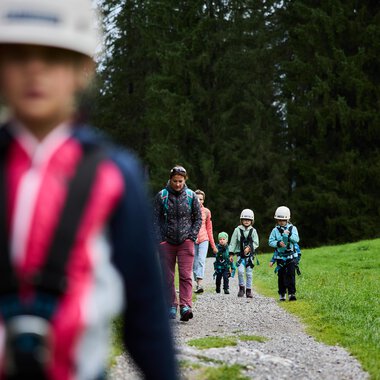Eine Gruppe von Kindern wandert auf einem Schotterweg durch einen Wald. Sie tragen Helme und sind gut ausgestattet für ein Abenteuer in der Natur. | © Kleinwalsertal Tourismus | Oliver Farys