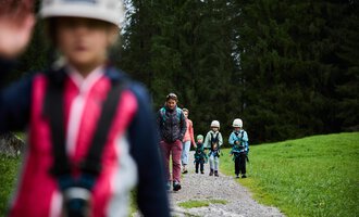 A group of children is hiking on a gravel path through a forest. They are wearing helmets and are well-equipped for an adventure in nature. | © Kleinwalsertal Tourismus | Oliver Farys