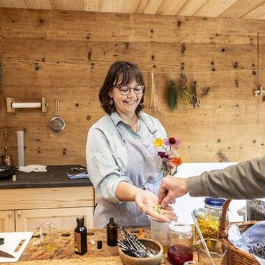 A smiling woman in a wooden kitchen is handing flowers and small bottles to a person. In the background, herbs and various ingredients are visible. | © Biohotel Walserstuba | Patrick Säly