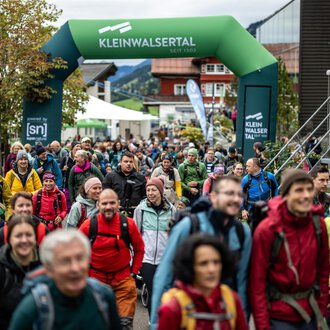 Zwei Wanderer gehen einen Pfad in den Bergen entlang. Die Sonne scheint und die Landschaft ist grün und goldfarben. | © Kleinwalsertal Tourismus eGen - Fotograf: Oliver Farys