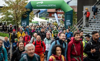 Zwei Wanderer gehen einen Pfad in den Bergen entlang. Die Sonne scheint und die Landschaft ist grün und goldfarben. | © Kleinwalsertal Tourismus eGen - Fotograf: Oliver Farys