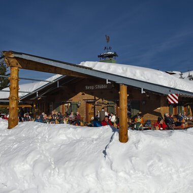 Eine gemütliche Berghütte im Schnee mit vielen Menschen auf der Terrasse. Die Umgebung ist winterlich und die Sonne scheint am blauen Himmel. | © Bergstüble | Michaela Beck Dornach