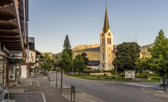 A quiet street with a small village and a church in the foreground. In the background, green trees and mountains can be seen. | © Kleinwalsertal Tourismus | Steffen Berschin