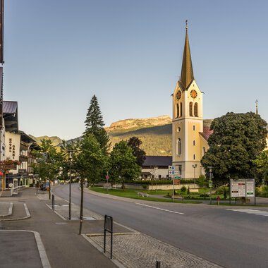 A quiet street with a small village and a church in the foreground. In the background, green trees and mountains can be seen. | © Kleinwalsertal Tourismus | Steffen Berschin