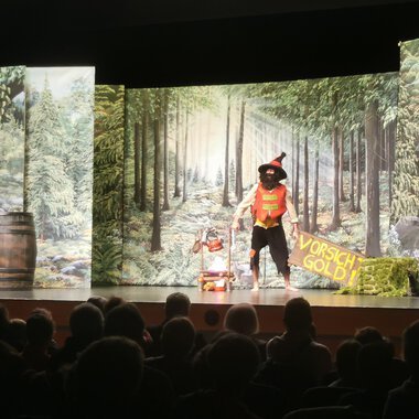 Eine Theateraufführung mit einem bunten Bühnenbild, das einen Wald zeigt. Ein Darsteller in einem Kostüm interagiert mit dem Publikum. | © KiWi Kindertheater GbR