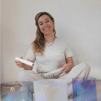 A smiling woman is sitting on the floor, holding a racket in her hand. In front of her are three colorful sound bowls. | © Genuss-Aktivhotel Sonnenburg | Anna Bader