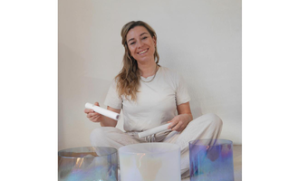 A smiling woman is sitting on the floor, holding a racket in her hand. In front of her are three colorful sound bowls. | © Genuss-Aktivhotel Sonnenburg | Anna Bader