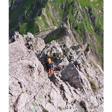 Eine beeindruckende Gebirgslandschaft mit steilen Felsen und grünen Wiesen. Ein Kletterer ist an einer Felswand unterwegs. | © stoamandl.at | Olli Sinz