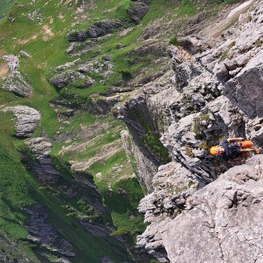 Eine beeindruckende Gebirgslandschaft mit steilen Felsen und grünen Wiesen. Ein Kletterer ist an einer Felswand unterwegs. | © stoamandl.at | Olli Sinz