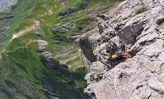 Eine beeindruckende Gebirgslandschaft mit steilen Felsen und grünen Wiesen. Ein Kletterer ist an einer Felswand unterwegs. | © stoamandl.at | Olli Sinz