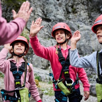 Three children in climbing gear greet an adult. They are wearing helmets and smiling against a rocky backdrop. | © Kleinwalsertal Tourismus | Oliver Farys