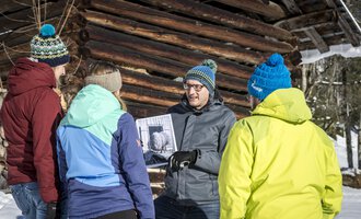 Eine Gruppe von fünf Personen steht im Schnee vor einer Holzscheune. Sie betrachten gemeinsam ein Foto, das einer der Teilnehmer in der Hand hält. | © Kleinwalsertal Tourismus | Dominik Berchtold