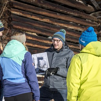 Eine Gruppe von fünf Personen steht im Schnee vor einer Holzscheune. Sie betrachten gemeinsam ein Foto, das einer der Teilnehmer in der Hand hält. | © Kleinwalsertal Tourismus | Dominik Berchtold